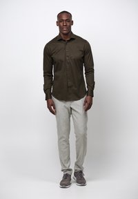 Mörk olivgrön knäppt skjorta, ljusgrå mönstrade byxor och grå sportskor, stående mot en enhetlig bakgrund.