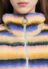 Fleecejakke med høj krave, der har horisontale striber i gul, pink, grå og blå. Guldlynlåsdetalje foran.