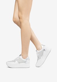 Sneaker platform bianche con accenti argentati, materiale in rete e design con lacci. Presentano una superficie testurizzata e suole lisce.