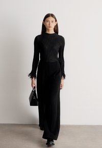 Žena kráčajúca v interiéri oblečená v kompletne čiernom outfite s dlhým rukávom a rebrovaným topom s perovými manžetami, širokými nohavicami, čiernymi topánkami a držiaca kabelku.