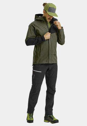 Water-resistant green jacket with black sleeves, zippered front pocket, and matching green cap, paired with black pants and hiking shoes.