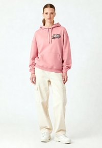 Hoodie rosa con "Amour" ricamato in nero, abbinato a pantaloni cargo crema con tasche laterali. Sneakers bianche chunky completano l'outfit.