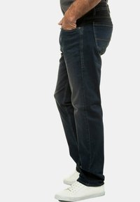 Tamsiai mėlynos džinsinės kelnės su tiesiomis kojomis, turinčios priekinius kišenes ir matomus siuvimo elementus. Dėvimos su baltomis sportinėmis bateliais.