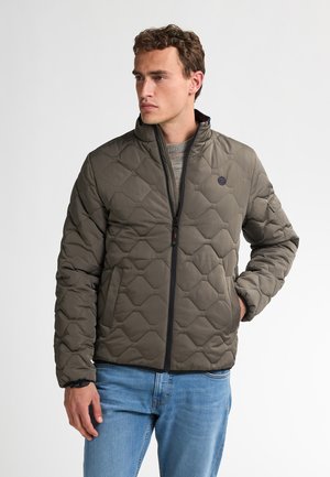 Man in een olijfgroene gewatteerde jas met zwarte ritssluiting en blauwe jeans, staand met één hand in zijn zak, kijkend naar links.