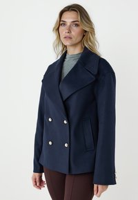 Cappotto doppiopetto blu navy in tessuto morbido, con grandi revers, bottoni dorati, tasche laterali e silhouette strutturata.