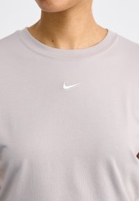 Šviesiai pilkas medvilninis marškinėlis su apvalia iškirpte, ant krūtinės srities esantis mažas baltas Nike logotipas ir švelni, minkšta tekstūra.