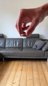 Anello in metallo con un design a catena d'argento tenuto in mano, sopra un divano in pelle grigia e un pavimento in legno. Lo sfondo presenta un cuscino grigio piegato.