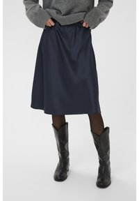Jupe A-line en satin marine avec une taille élastique, associée à des bottes en cuir noires montantes et un pull gris avec des poignets côtelés.