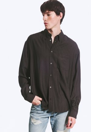 Jeune homme portant une chemise foncée ample à boutons et un jean bleu clair déchiré, regardant sur le côté avec une main dans la poche.