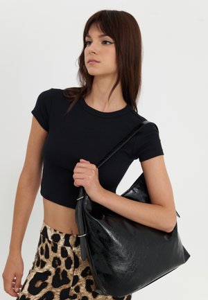 Jeune femme portant un crop top noir et une jupe imprimé léopard, tenant un grand sac à bandoulière noir sur l'épaule droite.