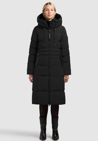 Sieviete, kas valkā garu melnu pūkaino mēteli ar kapuci, ar aizdari priekšpusei, sānu kabatām, melnām zeķubiksēm un melnām potīšu zoodēm uz pelēka fona.