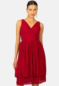 Robe plissée rouge avec un décolleté en V profond, un corsage ajusté et une jupe à plusieurs couches. Fabriquée en tissu léger, elle présente des plis verticaux et une texture lisse.