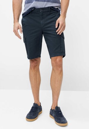 Mann trägt marineblaue Cargoshorts und marineblaue Sneakers mit Gummisohlen, steht vor einem schlichten weißen Hintergrund.