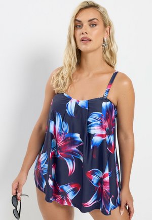 Blondes Frau mit blauem, ärmellosem, geblümtem Kleid, die schwarze Sonnenbrillen hält und vor einem weißen Hintergrund steht.