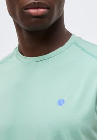 Κοντινό πλάνο του κάτω μέρους του προσώπου και του λαιμού ενός ατόμου που φοράει ένα ανοιχτό πράσινο πουκάμισο με ένα μικρό μπλε κοντομάνικο λογότυπο σε σχήμα ημισελήνου στο στήθος.