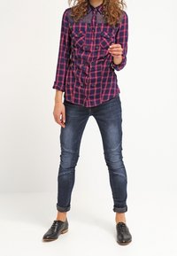 Chemise à carreaux en bleu marine et rose avec poches poitrine et section supérieure transparente, associée à un jean slim bleu foncé et des chaussures noires à lacets.