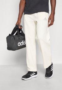 Mala de duffel preta com logotipo branco, calças texturizadas off-white, e sapatilhas de atletismo pretas com riscas brancas. Design simples, estilo funcional.
