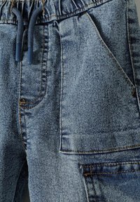 Denimske hlače s svetlo modrim opranjem, ki imajo elastičen pas, šnuro in več žepov z vidnimi šivi.