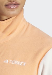 Sudadera de forro polar melocotón con cuello alto, que presenta un tejido de acento blanco y el logo bordado "TERREX" en el pecho. Textura suave.