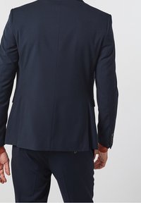 Marinblå skräddarsydd kavaj med strukturerade axlar, enkel sprund och knäppte manschetter; slät tyg med subtil lyster och snygga sömmar.