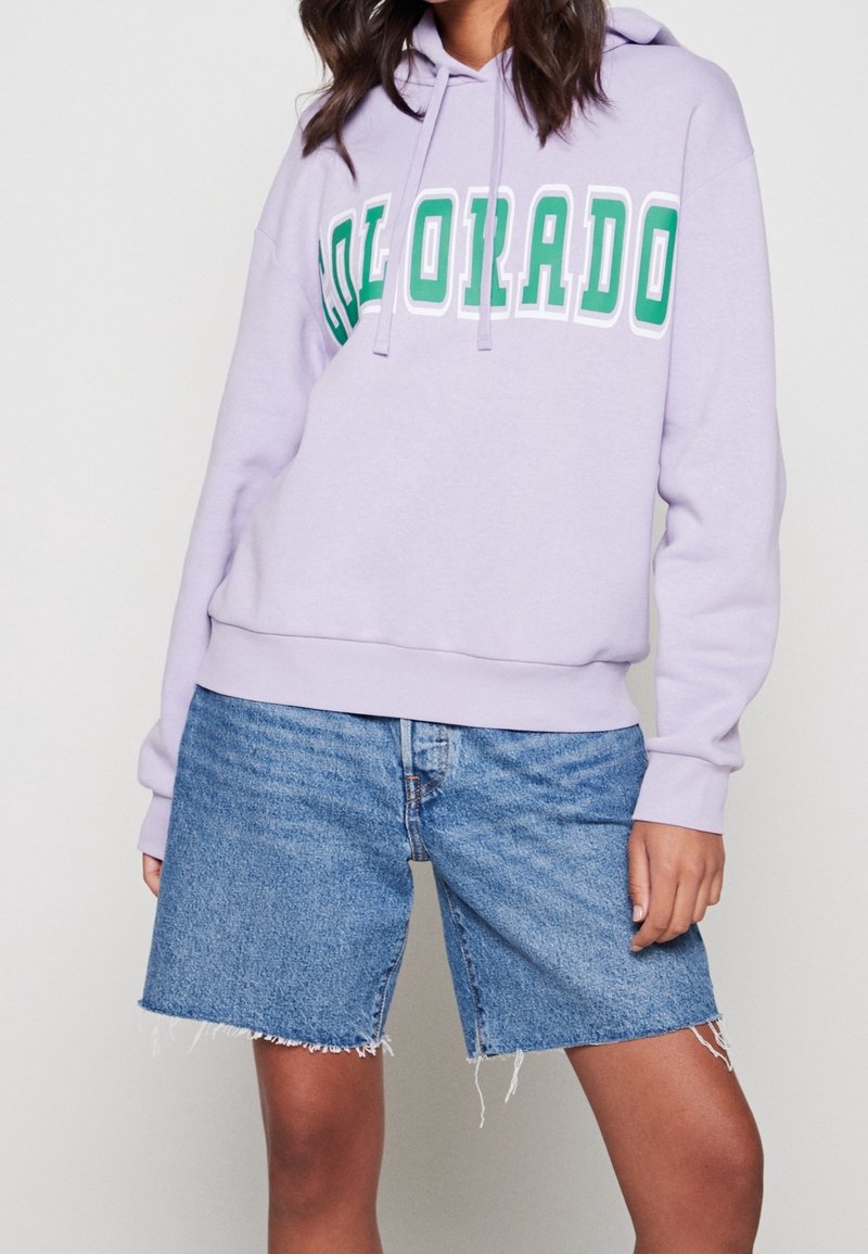 Persona che indossa un felpa leggera di colore viola con la scritta "COLORADO" in lettere verdi e jeans corti sfilacciati fino al ginocchio, in piedi su uno sfondo bianco.