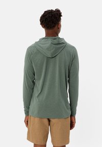 Maglietta a maniche lunghe verde con cappuccio, dal taglio comodo, con maniche raglan e una texture morbida e liscia. Abbinata a pantaloni corti beige.