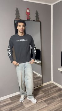 Zwart shirt met lange mouwen en witte vuurmotieven op de mouwen, gecombineerd met lichtblauwe wijde spijkerbroek en grijze sneakers. Spiegel en neutrale muren op de achtergrond.