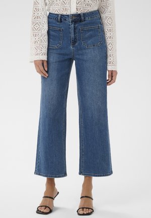 Femme portant un jean bleu taille haute à jambes larges, un chemisier blanc en crochet à manches longues et des talons ouverts noirs à brides, debout sur un fond blanc.