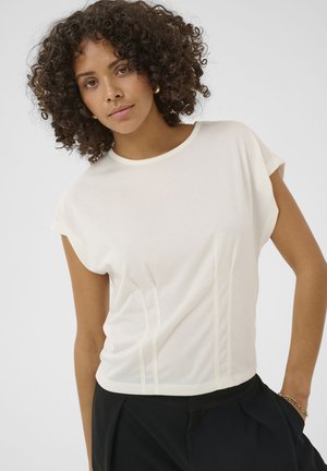 Kobieta o kręconych włosach, ubrana w biały krótki rękaw i czarne spodnie, stojąca z jedną ręką w kieszeni na jednolitym tle.