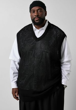 Man met baard die een zwarte gebreide muts draagt, een zwart mesh sweatervest over een wit overhemd, zwarte broek en een zilveren horloge en ringen, staand tegen een grijze achtergrond.