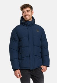 Uśmiechnięty mężczyzna w granatowej ocieplanej kurtce z kapturem, stojący z jedną ręką w kieszeni na jednolitym tle.