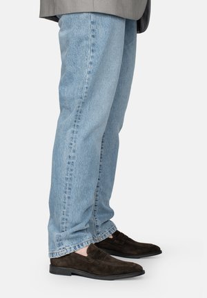 Person trägt hellblaue Jeans und dunkelbraune Wildlederloafers und steht vor einem schlichten weißen Hintergrund.