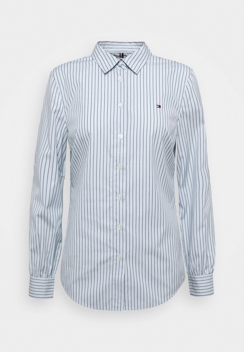 Chemise blanche à manches longues avec boutons, fines rayures verticales noires et petit logo sur la poitrine gauche.