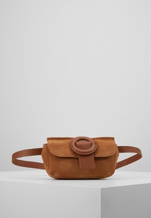 Riñonera de ante marrón con una correa de cuero y hebilla circular de cuero, exhibida sobre un pedestal blanco contra un fondo gris liso.