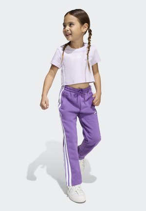 Ung pige med fletninger iført en lyserød crop top, lilla joggingbukser med hvide striber og hvide sneakers går og smiler.