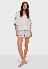Ensemble de pyjama à manches courtes, couleur crème clair, composé d'un haut à boutons et d'un short assorti. Présente un motif subtil et une finition noire le long des bords.