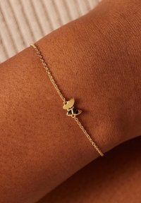 Gold chain bracelet featuring a small heart and bee charm. The bracelet is delicate, textured, and sits on a brown skin tone.