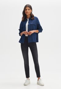 Dark blue denim jacket with two chest pockets, paired with a white t-shirt and black jeans. Model wears white sneakers. Plain background.