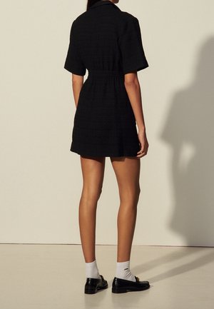 Kobieta stojąca tyłem do kamery, ubrana w krótką czarną sukienkę, białe skarpetki i czarne mokasyny na jednolitym beżowym tle.