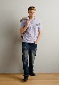Lilla t-skjorte med trykk på forsiden, sammen med blå denim jeans og svarte sko. Modellen bærer en beige ryggsekk og står på tregulv.