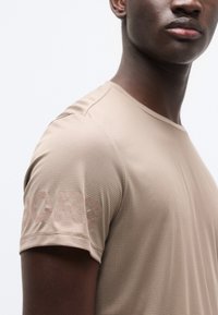 Μπεζ αθλητικό μπλουζάκι με υφασμάτινο μοτίβο και κοντά μανίκια, με διακριτικό λογότυπο στον ώμο. Λείο ύφασμα, εφαρμοστή σχεδίαση.