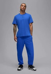 T-shirt in cotone blu con scollatura rotonda e maniche corte, abbinato a pantaloni jogger blu coordinati con un piccolo logo. Scarpe da ginnastica nere.