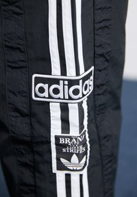 Melnas sporta bikses ar baltām vertikālām svītrām, izsistītu "adidas" logo un uzdoto emblēmu ar brenda ikonisko trifeli.