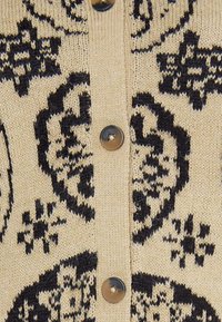 Stickad cardigan i beige med svart mönster. Har runda svarta knappar, texturerad tyg och intrikata motiv genom hela designen.
