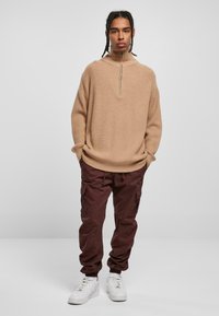 Beige ribbestrikket genser med halvglidelås, sammen med bordeaux cargobukser som har sidelommer og elastiske mansjetter, fullført med hvite sneakers.