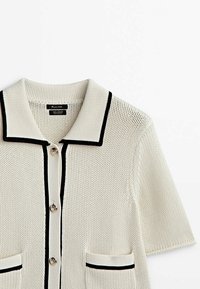 Sweter rozpinany z kremowej bawełny z czarnym wykończeniem. Posiada kołnierzyk, krótkie rękawy, dwie przednie kieszenie oraz okrągłe guziki. Teksturowana powierzchnia.