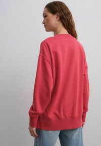 Różowy, oversize'owy sweter z żebrowanymi mankietami i dołem. Miękka tekstura i okrągły dekolt, w zestawieniu z jasnoniebieskimi dżinsami.