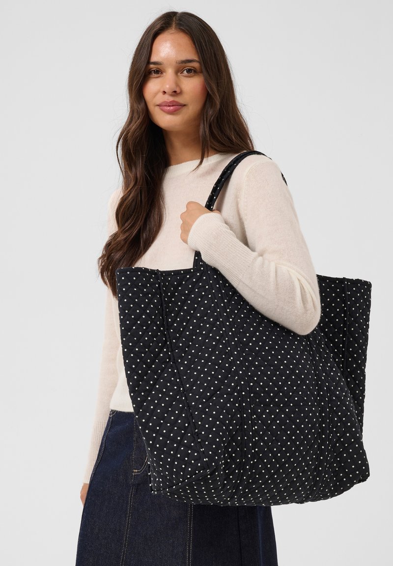 Femme aux longs cheveux bruns portant un pull beige et une jupe foncée, tenant un grand sac cabas noir matelassé avec des pois blancs.