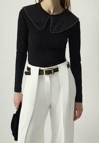 Mujer vistiendo una blusa negra de manga larga con gran cuello y pantalones blancos de cintura alta, sosteniendo un accesorio negro en su mano izquierda.