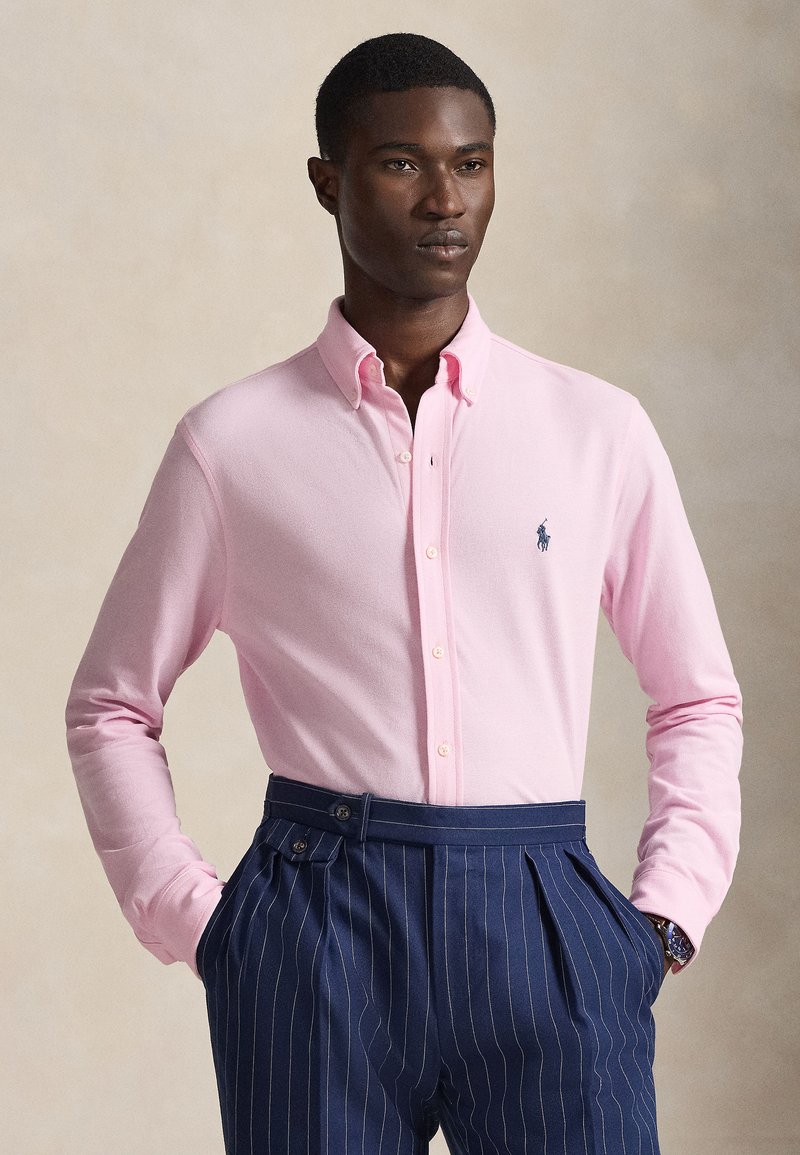 Light pink long-sleeve shirt with button-down collar, featuring a small navy logo on the chest. Paired with navy pinstriped trousers.